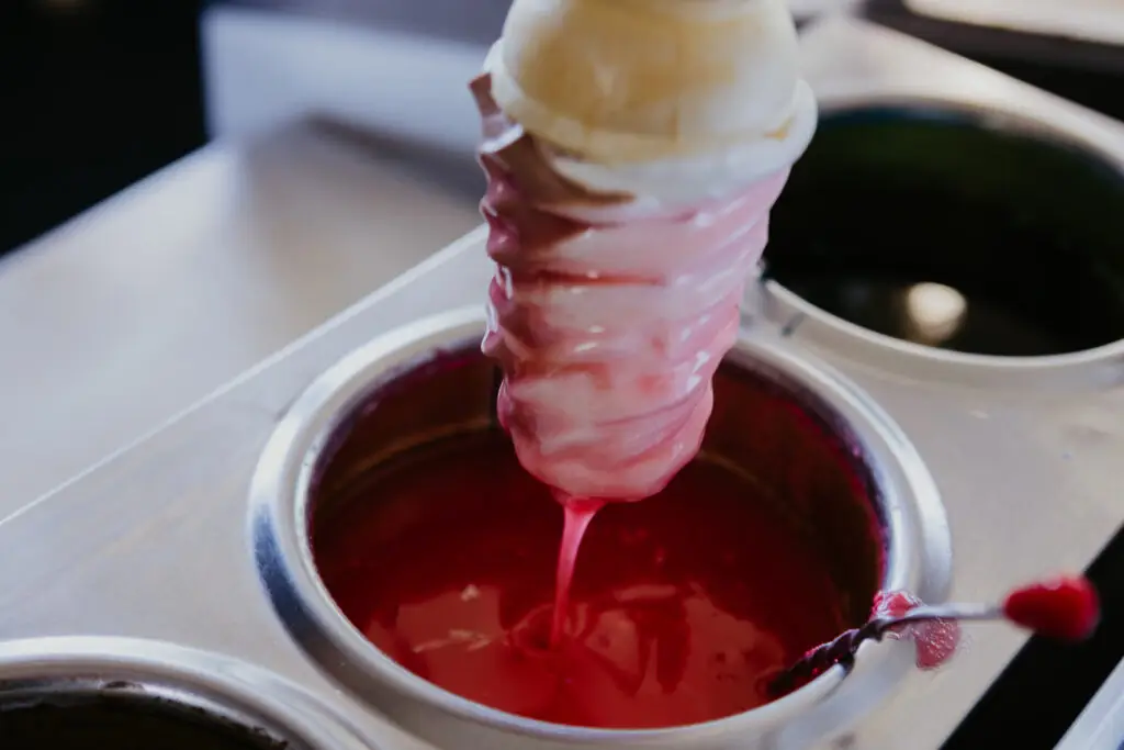 A vanilla ice cream cone being dipped into bright red cherry coating, creating a colorful candy-covered treat at Buoy 18.