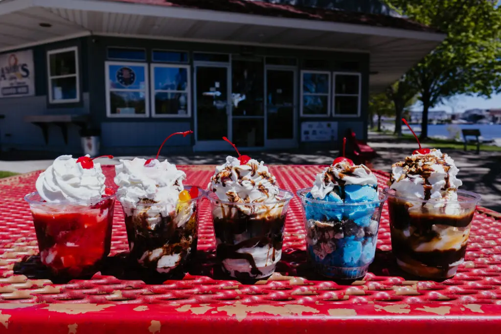 Five ice cream sundaes topped with whipped cream, chocolate sauce, and cherries displayed on a red picnic table outside Buoy 18.